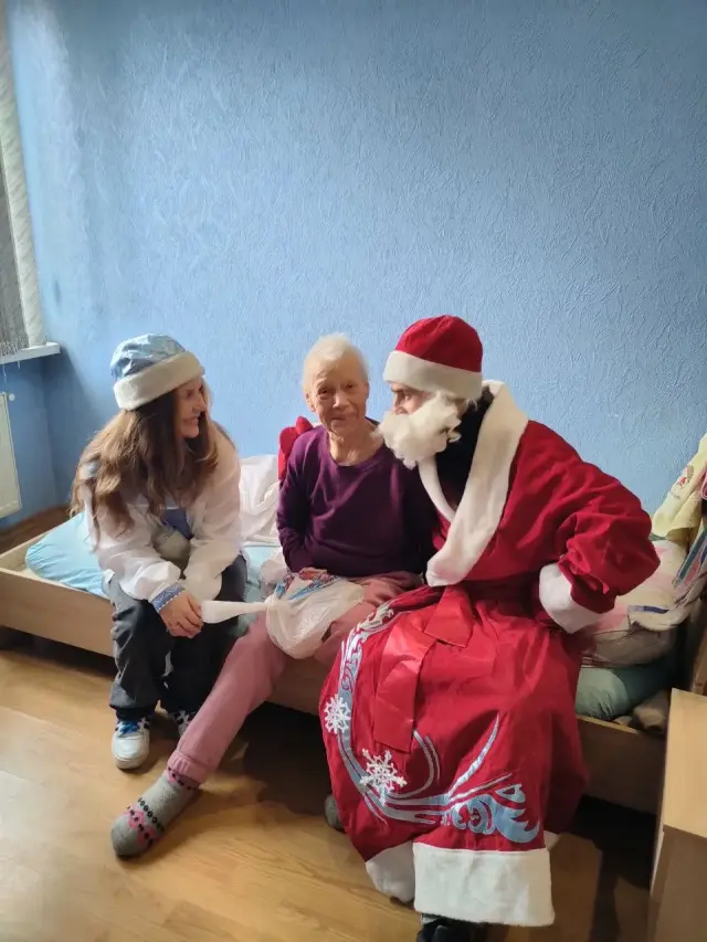 Аніматори у костюмах Діда Мороза та Снігуроньки сидять поруч із літньою жінкою на ліжку в кімнаті пансіонату та вручають їй пакунок із подарунком. Кімната зі світло-блакитними стінами, дерев'яною підлогою та ліжками.