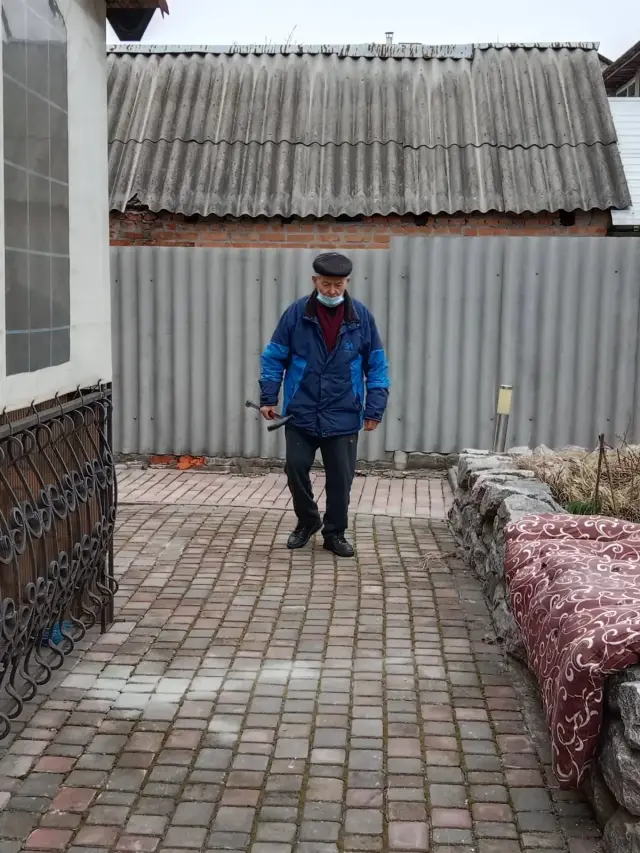 Літній чоловік у кепці та синій куртці прогулюється з паличкою по викладеному бруківкою подвір'ю. Навколо огорожа, кам'яна клумба та кована решітка біля будівлі.