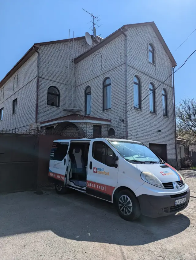 Білий мікроавтобус служби медичного транспортування з відчиненими бічними дверима припаркований перед триповерховою будівлею з білої цегли з арковими вікнами.