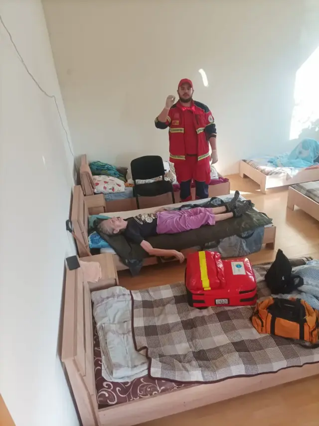 Парамедик у червоній формі та кепці стоїть у кімнаті з кількома ліжками поруч із літньою жінкою, яка лежить на м'яких транспортувальних ношах; поряд червоний медичний рюкзак і сумка з обладнанням.