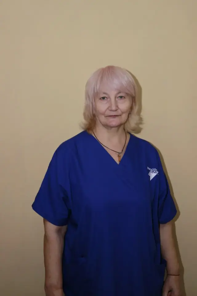 Жінка зі світлим волоссям у темно-синьому медичному костюмі з логотипом Health ONE стоїть біля бежевої стіни. Портретне фото медичної працівниці пансіонату.