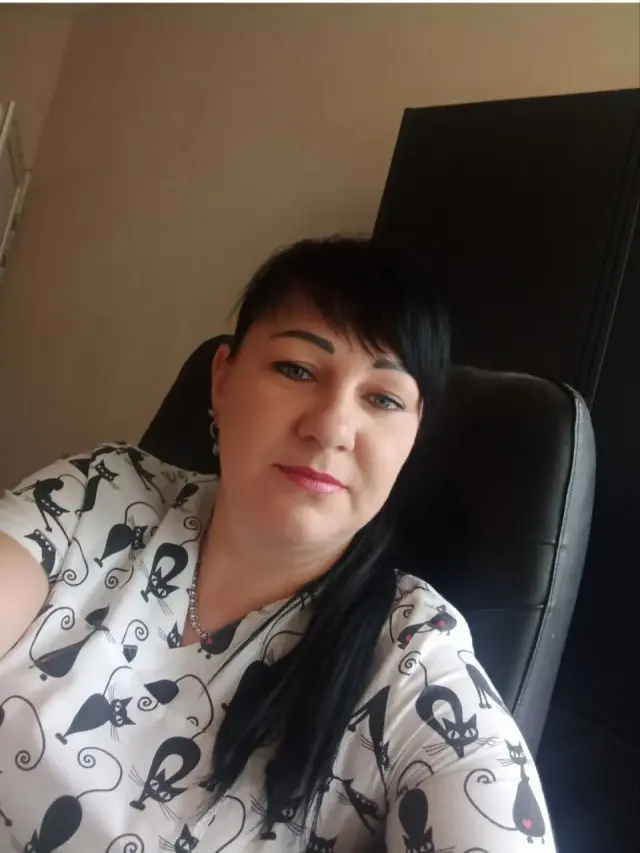 Жінка середнього віку з темним волоссям у світлій блузі з принтом котиків сидить у чорному офісному кріслі в кабінеті. Портретне фото співробітниці закладу.