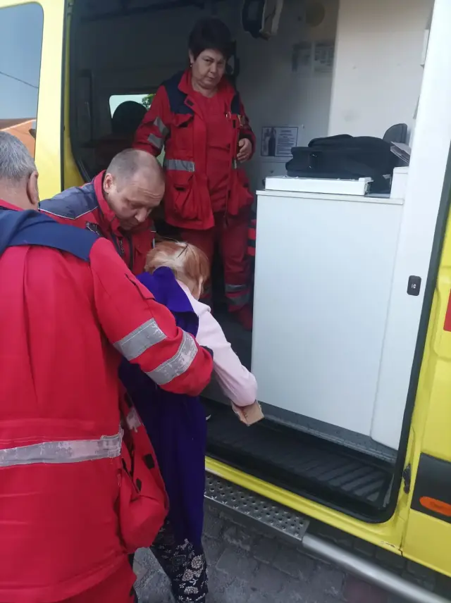 Медики в червоних уніформах допомагають літній жінці в бузковому одязі піднятися сходинкою до жовтого автомобіля швидкої допомоги. Фельдшерка стоїть усередині салона і приймає пацієнтку.