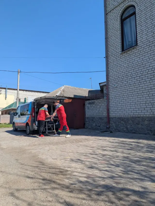 Двоє працівників медичного транспорту в червоній формі вивантажують ноші-каталку з мікроавтобуса на брукованому подвір'ї. На задньому плані — цегляна будівля та ворота.